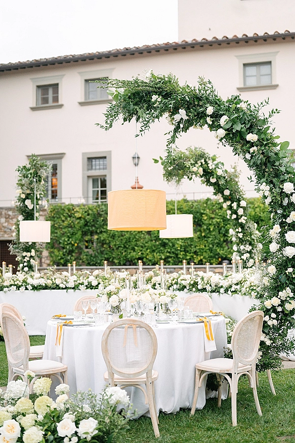 Reception tablescape with a round wedding sweetheart table, white roses and greenery garland, taper candles, and pendant lights on a lawn by a stucco building
