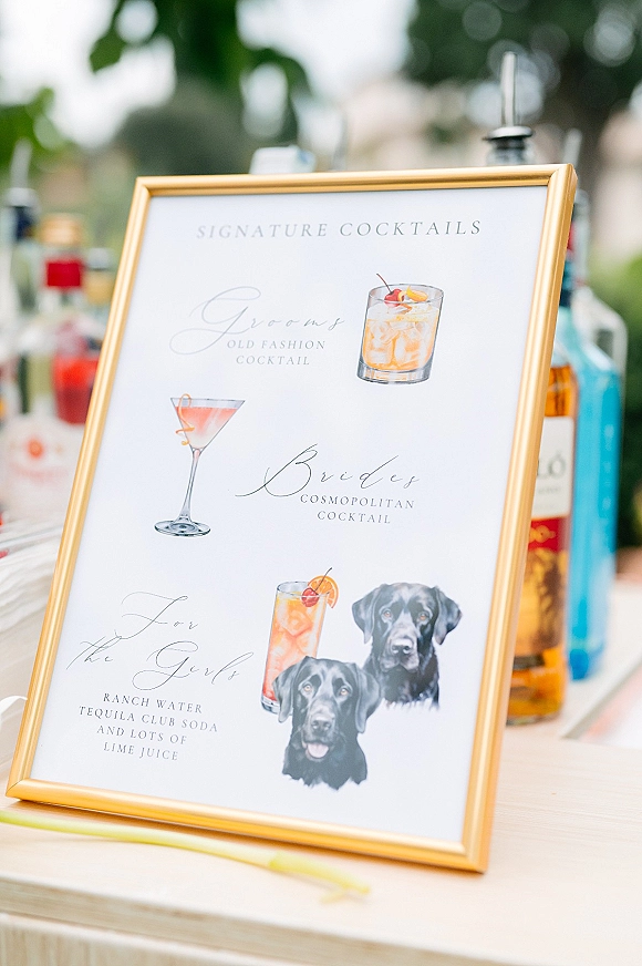 Signature cocktail sign in a gold frame with illustrated drink menu on a bar table beside liquor bottles, set against soft outdoor greenery bokeh