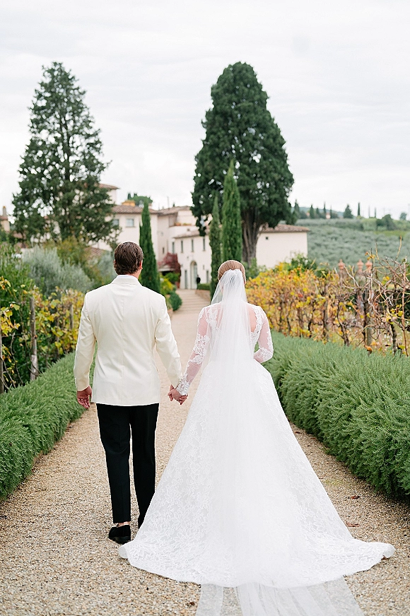 Couple portrait of newlyweds holding hands walking away on a gravel garden path, bride’s veil and long train beside groom in white jacket