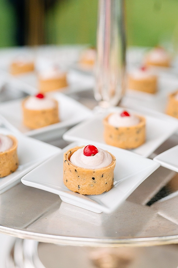Wedding dessert table with mini tartlets on white square plates, whipped cream and red berry garnish on a tiered stand outdoors