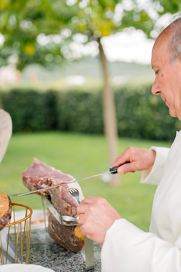 Wedding catering station with a ham carving station featuring a cured ham leg on a stand with carving knife and fork on a white tablecloth outdoors