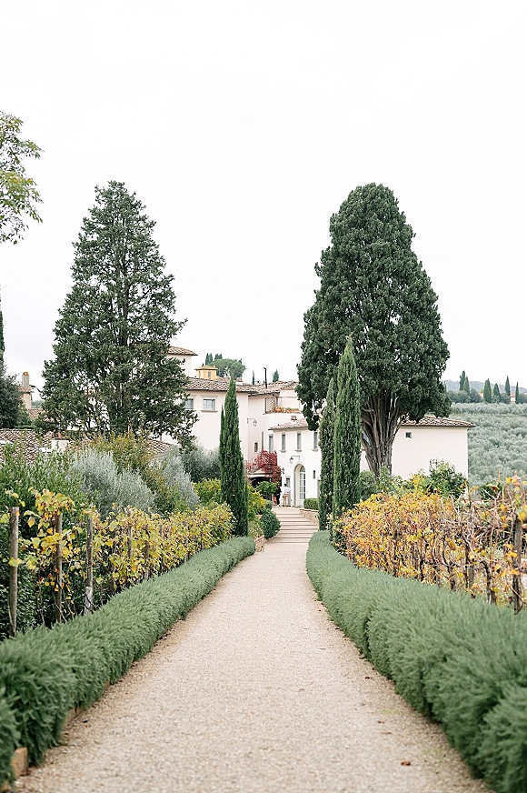 Wedding venue exterior with a gravel driveway leading to a white villa, framed by hedges and trees with vineyard rows and hills beyond