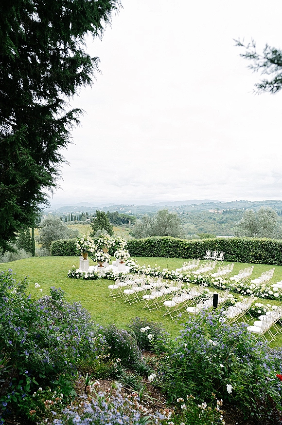 Ceremony setup with white folding chairs and white rose aisle florals arranged on a lawn, framed by hedges and rolling hills under clouds