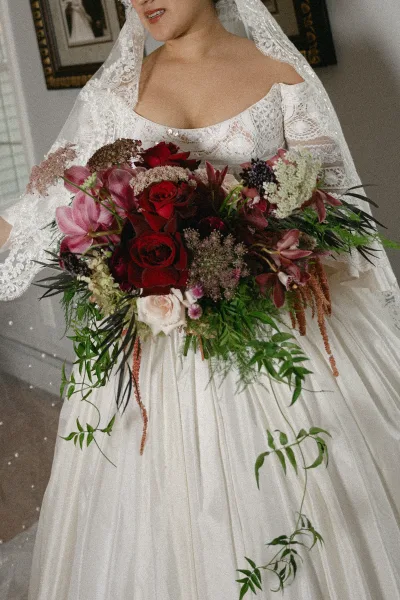 Bridal bouquet of red roses and pink blooms with trailing greenery, held against an off-the-shoulder lace dress and veil indoors.