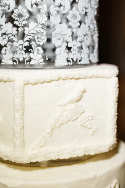 Wedding cake with white buttercream frosting, piped border, and embossed damask stencil design, set against a simple brown backdrop