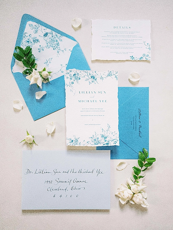 Invitation suite with blue wedding invitations on a light gray surface, deckled-edge cards with calligraphy, floral art and greenery sprigs