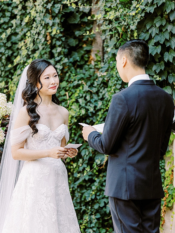 Wedding vows as groom reads from vow cards to veiled bride in off-the-shoulder lace dress, bouquet in hand, before an ivy wall