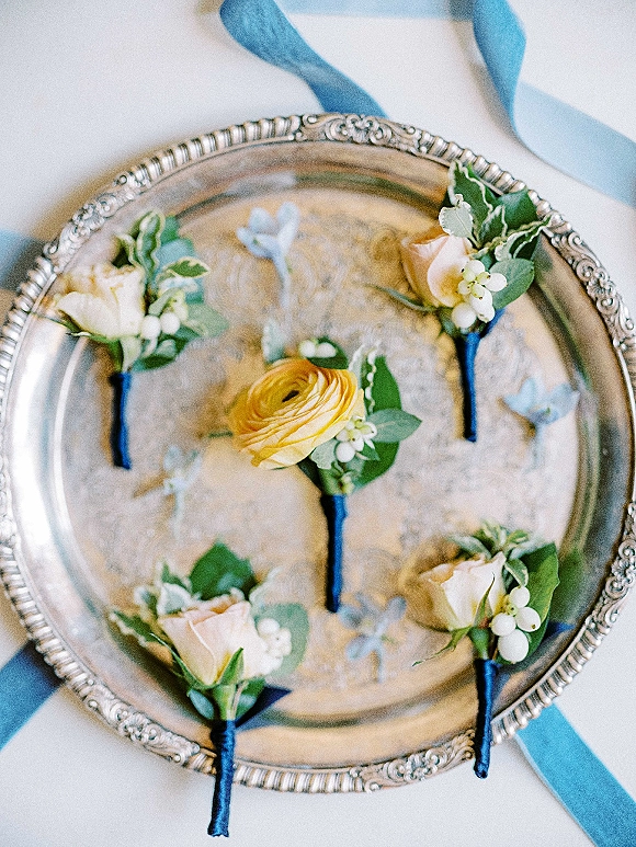Wedding boutonnieres with white roses, yellow ranunculus and greenery, wrapped in blue ribbon on a silver tray against white fabric backdrop