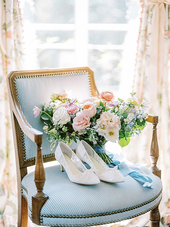 Bridal details featuring wedding bouquet and shoes, white lace heels and a bouquet tied with a blue ribbon by a window-lit chair