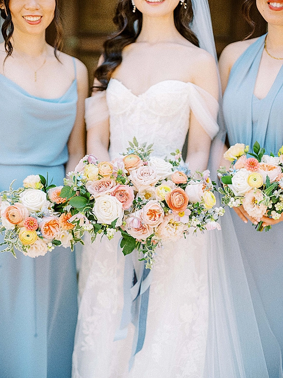 Bridesmaid group portrait with bride holding pastel floral bouquets and blue ribbon accents, bridesmaids in blue dresses by a blurred stone wall