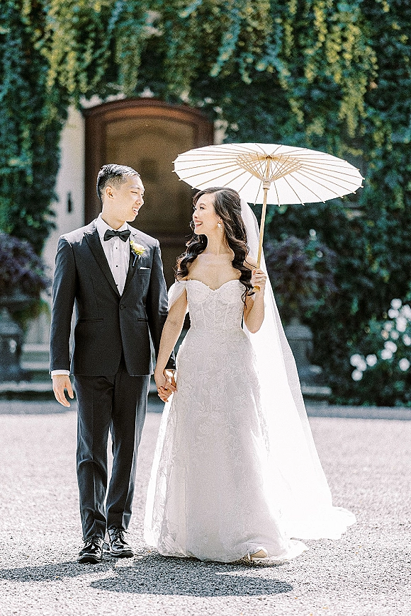 Couple portrait of bride and groom holding hands, she holds a white parasol in a sunlit ivy courtyard by an arched doorway