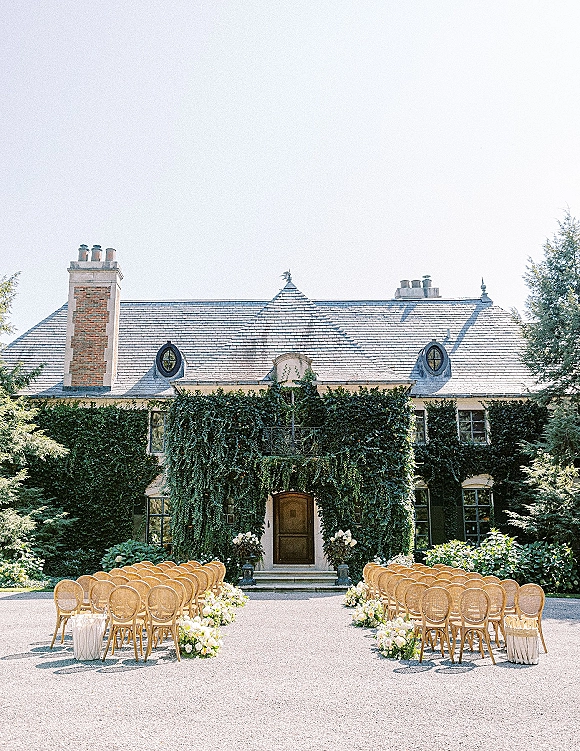 Ceremony setup with outdoor ceremony seating of rattan chairs and pastel aisle florals in a gravel courtyard before an ivy-covered house