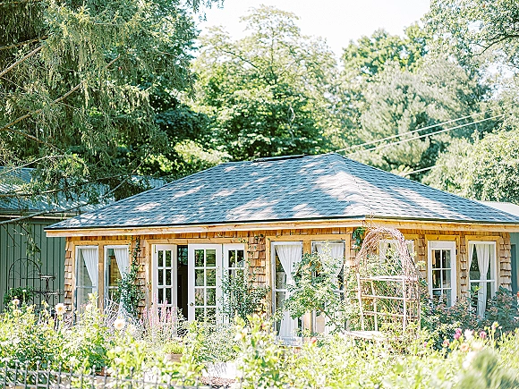 Garden wedding venue with an intimate wedding venue cottage, French doors and white curtains, climbing vines, and a bench by flower beds