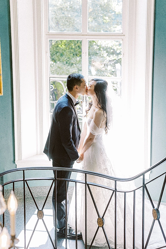 Wedding kiss portrait of bride and groom kissing by a large window, hands clasped, veil backlit, groom in tuxedo near staircase railing
