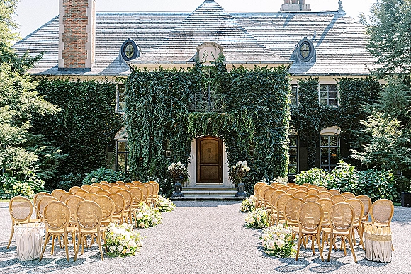 Ceremony setup with cane back chairs and pastel aisle florals lining a gravel courtyard before an ivy-covered building entrance