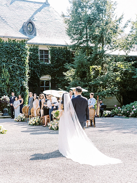 Processional moment as bride walking down aisle in a long veil and gown train, bouquet in hand, toward groom in a sunny ivy courtyard