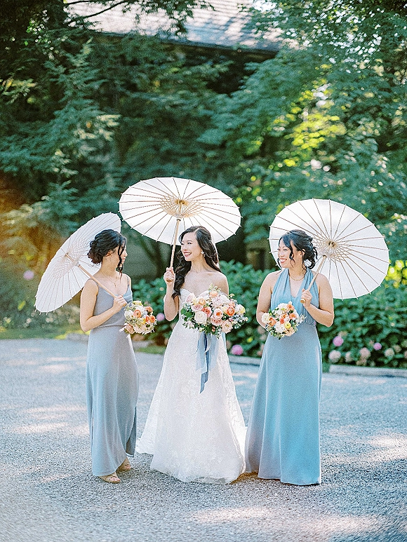 Bridesmaid photo of bride with bridesmaids holding paper parasols, carrying peach blush bouquets along a garden gravel driveway with trees