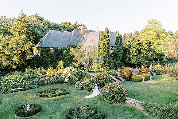 Couple portrait of bride and groom in garden, bride in wedding dress with cathedral veil holding bouquet by ivy-covered house steps