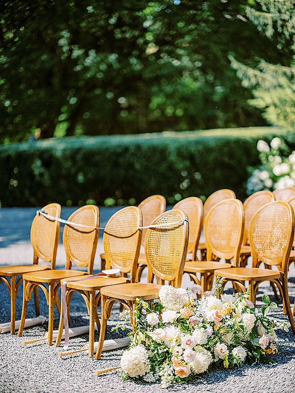 Ceremony seating with outdoor wedding chairs, cane-back wood rows lining a gravel aisle with rose and hydrangea ground florals in a garden setting