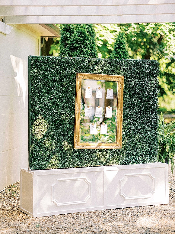 Seating chart display on a boxwood seating chart wall with gold frame and wax-sealed escort cards pinned above a white pedestal outdoors in sunlight