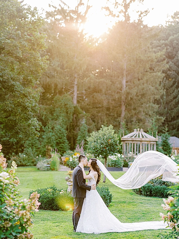 Wedding kiss portrait of bride and groom kissing, long cathedral veil blowing in wind on a garden lawn with gazebo at sunset
