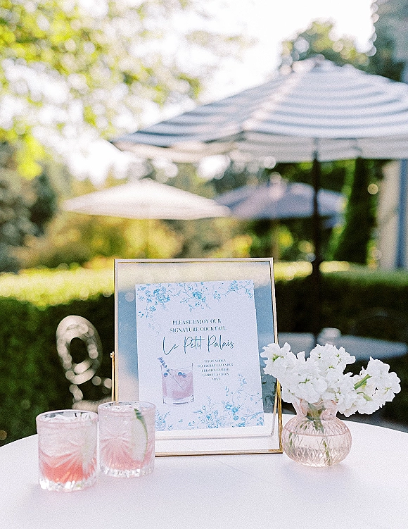 Signature cocktail sign with wedding signature cocktail menu in a glass frame beside cocktail glasses and white flowers on an outdoor patio table