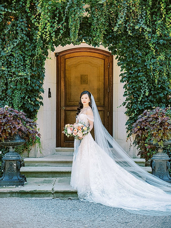 Bridal portrait of a bride holding bouquet in a strapless lace gown with cathedral veil on stone steps by a wooden door and ivy wall
