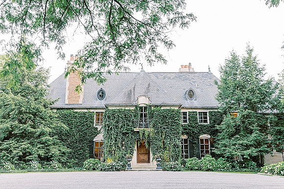 Wedding venue exterior with ivy and French doors on a stone manor house, balcony railing and lantern planters along a gravel driveway