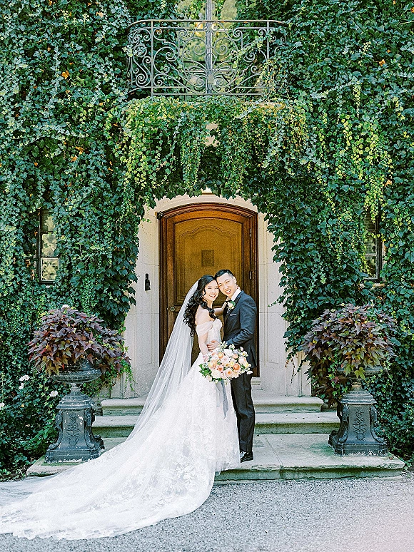 Couple portrait of bride and groom embrace, her long veil and bouquet flowing on stone steps before an ivy-covered wall and door