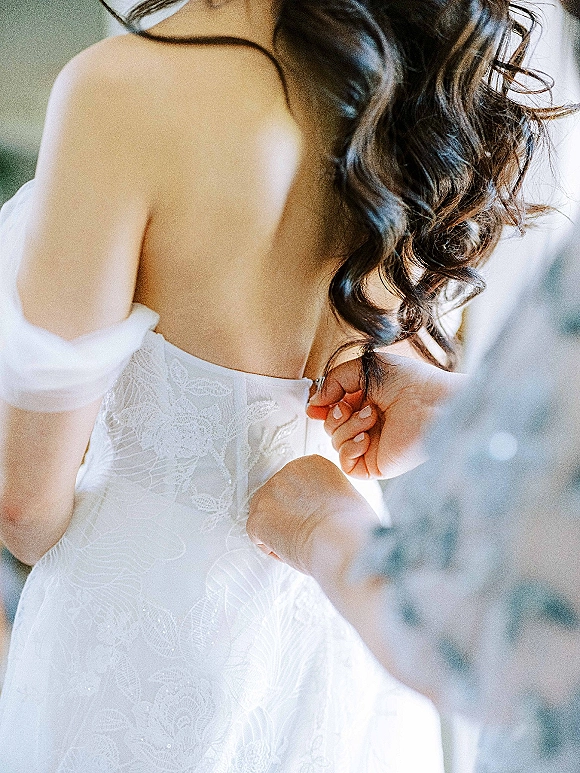Wedding dress details in a lace wedding dress with off-the-shoulder sleeves and back zipper, brunette curls in soft indoor light