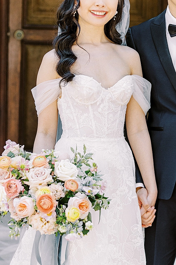 Wedding couple portrait of bride and groom holding hands, her pastel bouquet and veil beside his black tuxedo against a wooden door backdrop