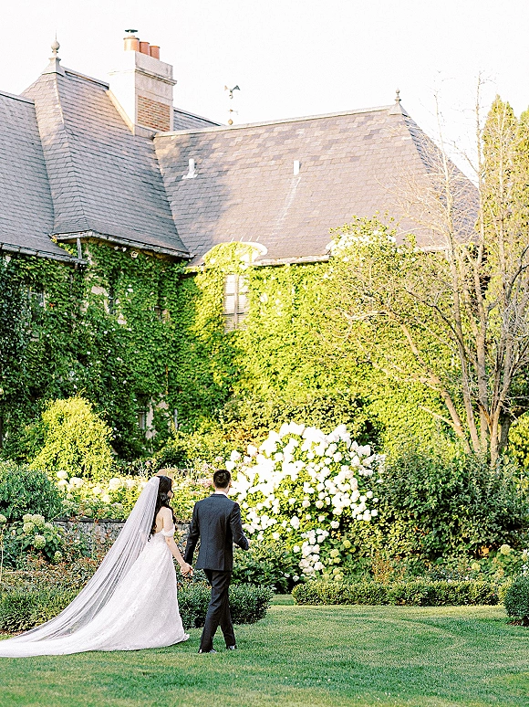 Couple portrait of bride and groom walking away holding hands, cathedral veil trailing over gown train in an ivy-covered garden estate