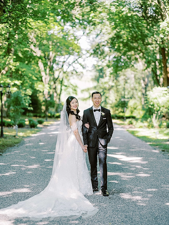 Couple portrait of bride and groom holding hands, her lace gown and long veil trailing as they walk a sunlit tree-lined road