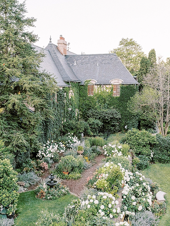 Garden wedding venue with a stone path lined by white hydrangeas leading to a fountain beside an ivy-covered house with shutters