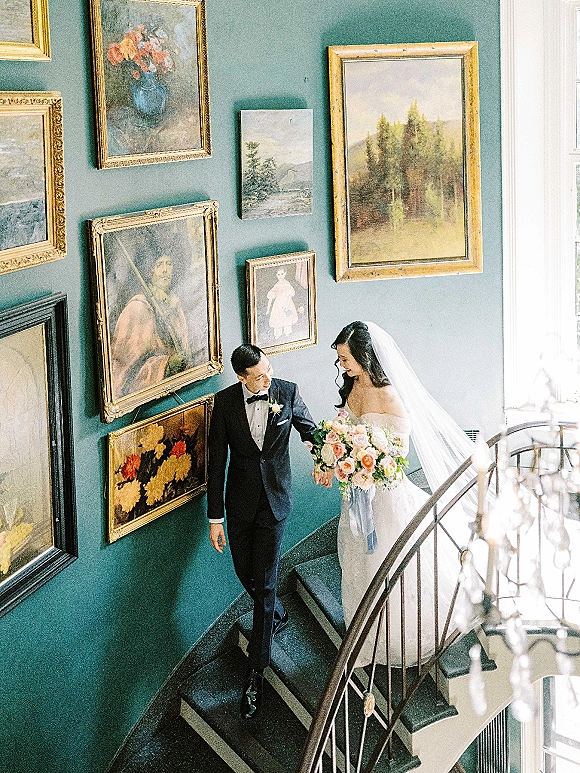 Couple portrait of bride and groom on staircase, holding hands as she holds a bouquet under chandelier crystals by a teal gallery wall