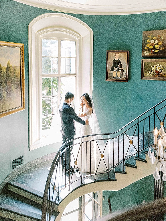 Couple portrait of bride and groom on staircase, holding hands in natural window light by arched window, veil glowing, chandelier above