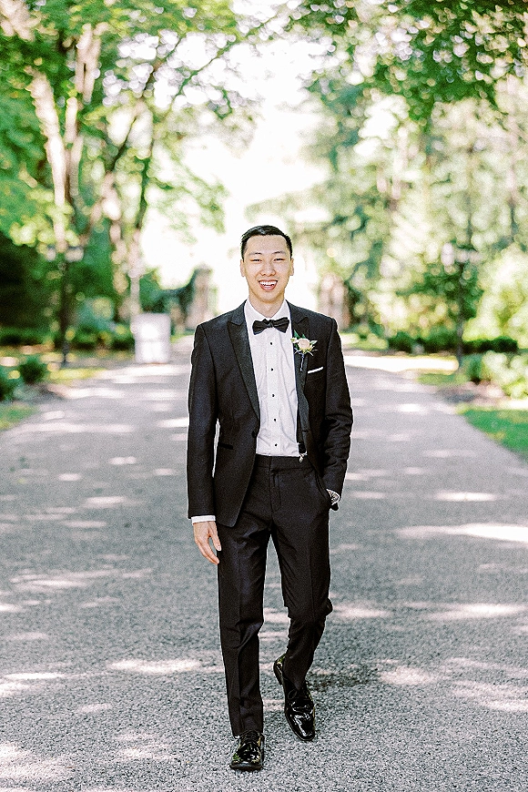 Groom portrait in a black tuxedo with bow tie and boutonniere, standing on a sunlit tree-lined road with greenery behind him