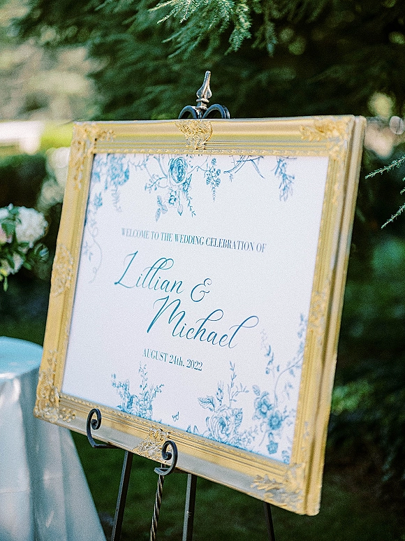 Wedding welcome sign in a gold ornate frame on an easel, with blue floral illustration, set against lush garden greenery outdoors