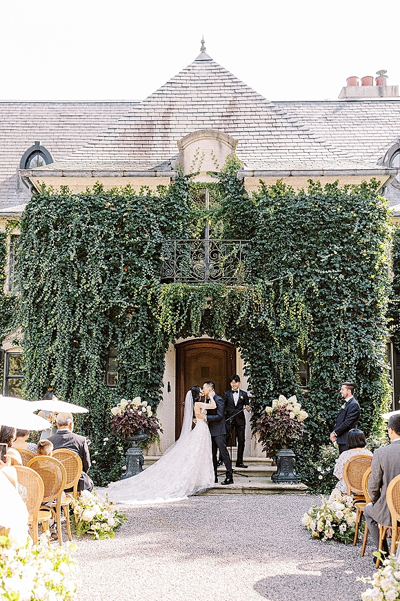 Ceremony kiss at outdoor wedding ceremony under a greenery arch, bride’s veil and train flowing as officiant stands before ivy estate doors
