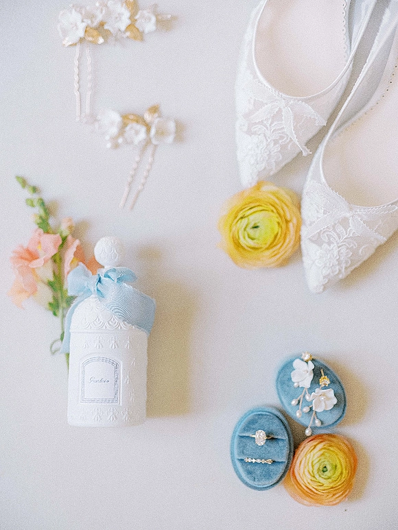 Wedding detail flatlay with wedding rings flatlay in a blue velvet ring box, lace bridal shoes, perfume, hair pins, flowers on white surface