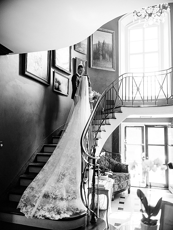 Wedding first look as groom seeing bride on a curved staircase, her long veil and lace train trailing down steps in bright entryway