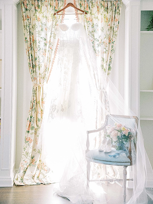 Wedding dress on hanger, strapless lace gown with tulle veil and blue ribbon by a bright window, bouquet and shoes nearby