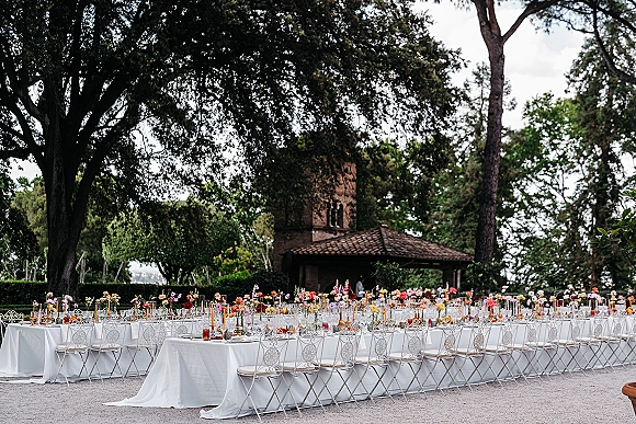 Reception tablescape for an outdoor wedding reception with long banquet tables, taper candles, colorful bud vases, and wrought iron chairs in a garden courtyard