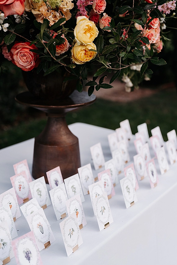 Wedding escort cards arranged in wooden card holders beside a rose and greenery centerpiece in a metal vase on an outdoor lawn table