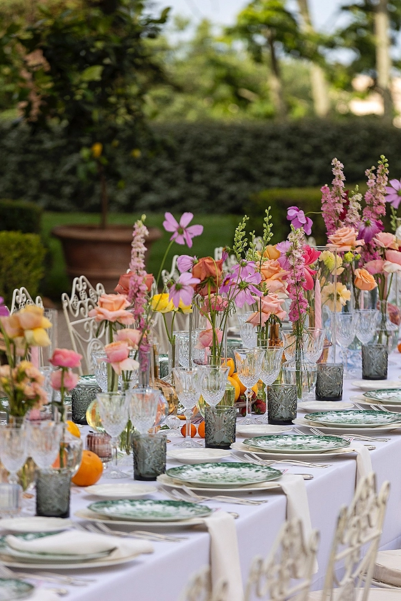 Reception tablescape with colorful bud vase centerpieces, crystal glasses, green patterned plates, and citrus accents on a long garden table