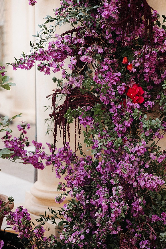 Wedding floral installation with cascading purple flowers and red blooms draped over stone columns against a building facade