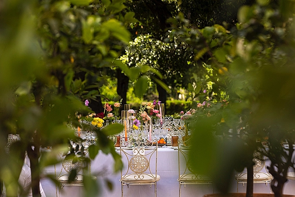 Reception tablescape with white linen, pastel floral centerpieces, taper candles in glass cylinders, and fruit bowl on long table under garden trees