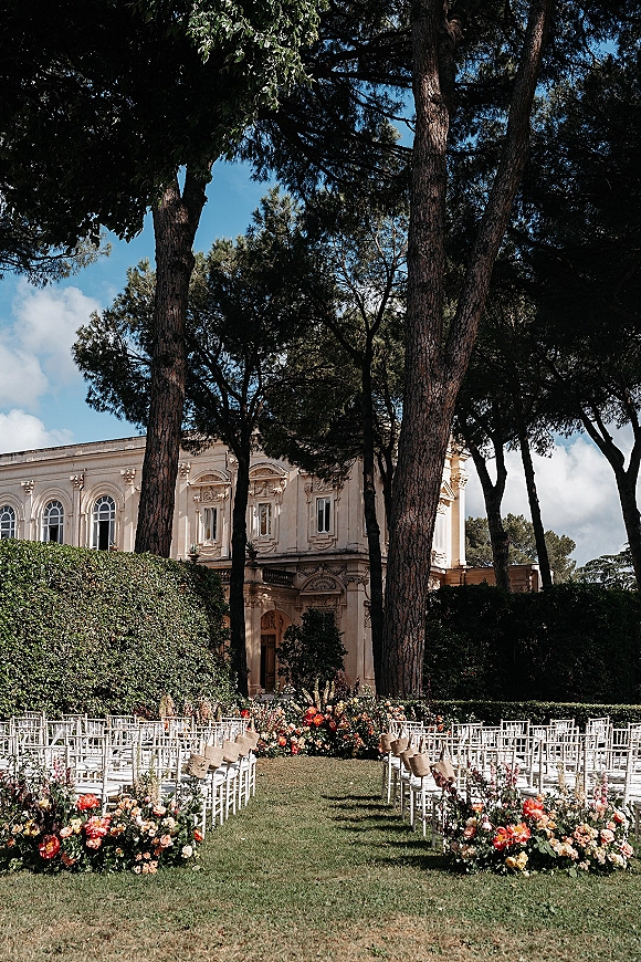 Ceremony setup with outdoor wedding ceremony setup, white chairs and floral-lined aisle on a lawn beneath tall trees by a historic building