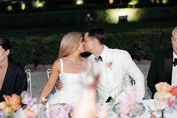 Wedding kiss as bride in strapless gown and groom in white tuxedo jacket lean in at candlelit sweetheart table in garden hedges