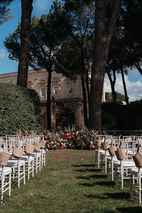 Ceremony setup with outdoor wedding ceremony setup featuring white chiavari chairs with straw bags on a lawn before a villa and pine trees